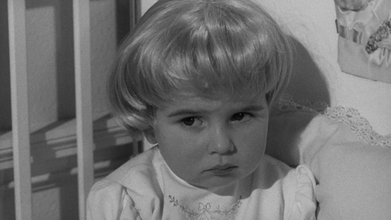 Village of the Damned (1960) movie still: closeup — A young child sits inside a white wooden crib structure with an embroidered shirt. The child…