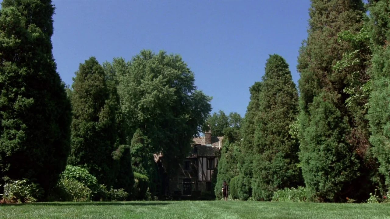 Creepshow (1982) movie still: establishing — A long green lawn is flanked by tall, dense coniferous trees on both sides. A large multi-story stone…