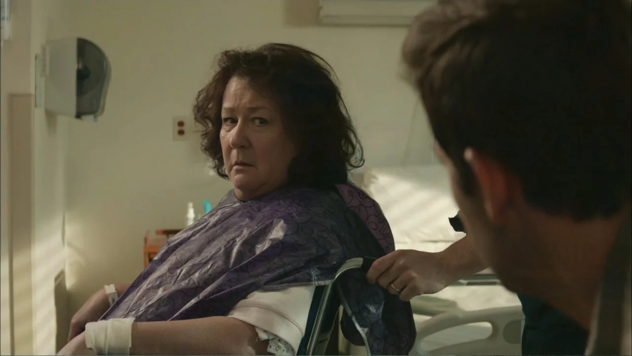The Hollars (2016) movie still: medium — A hospital room with a patient in a wheelchair and an attendant nearby. A man is pushing the wheelchair…