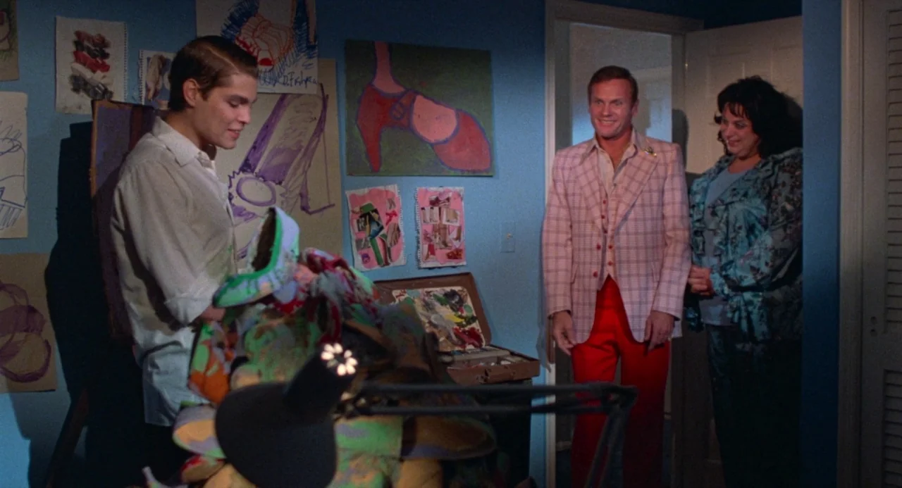 Polyester (1981) movie still: medium — A blue-walled room decorated with various colorful drawings of high-heeled shoes on paper. A young man shows…