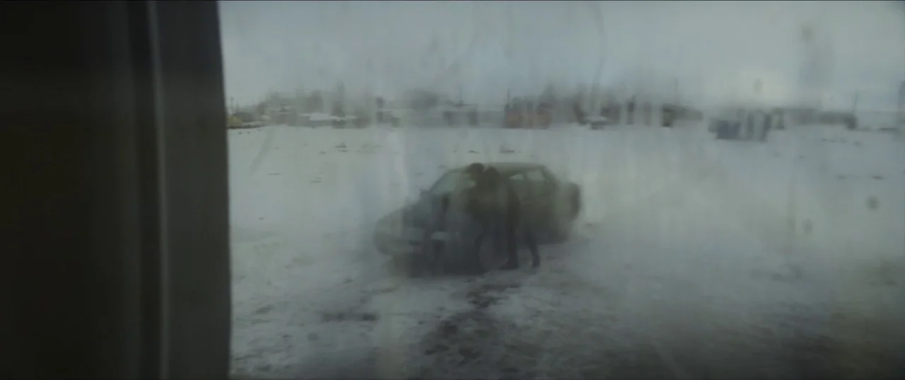 About Dry Grasses (2023) movie still: wide — A snowy, grey-toned outdoor area viewed through a condensation-covered pane of glass. Two indistinct…
