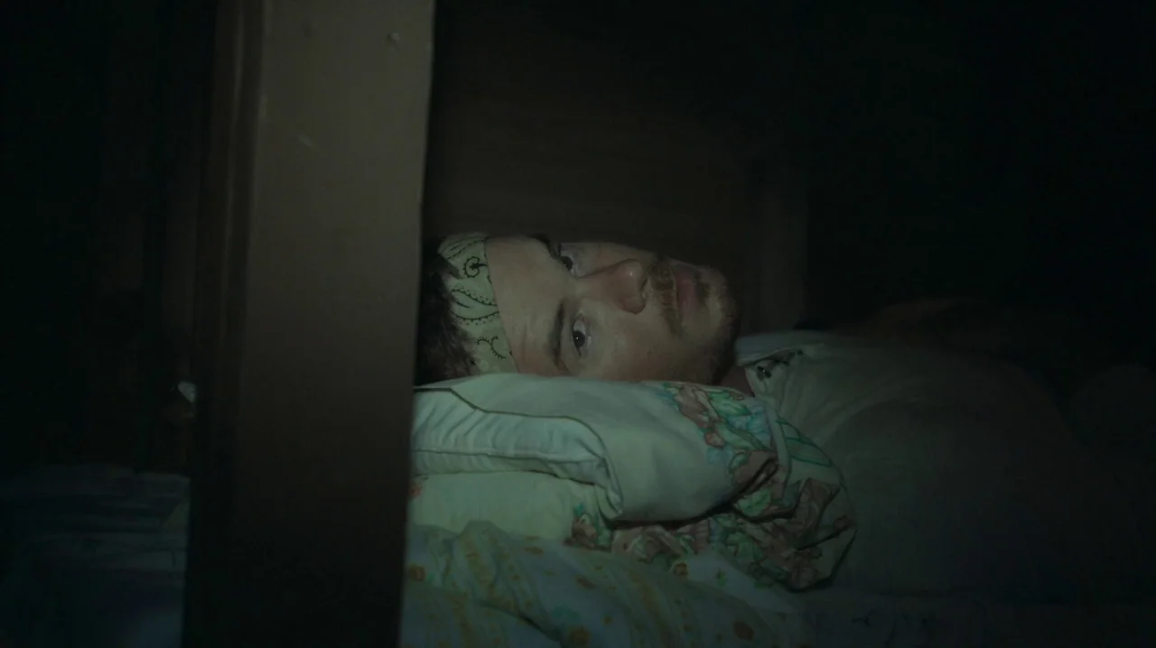The Sacrament (2013) movie still: closeup — A man lies on a pillow in a dark sleeping bunk bed. The person looks toward the camera while resting in…