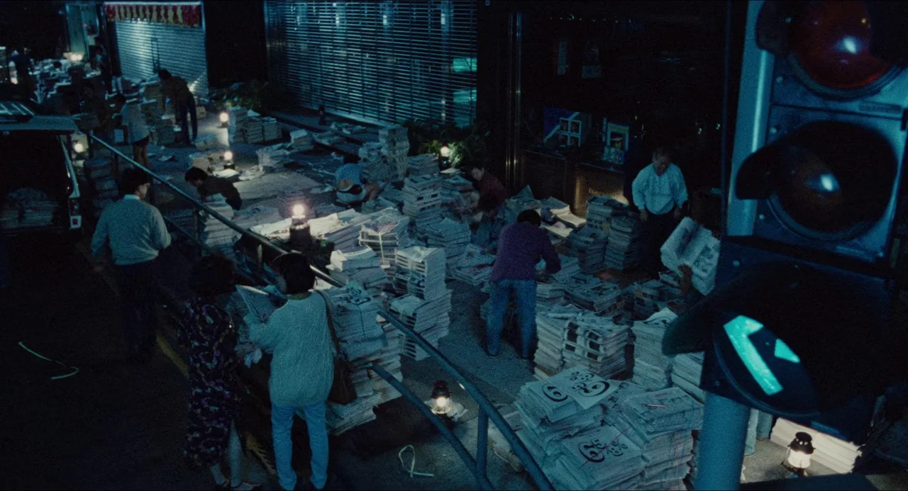 Rouge (1987) movie still: wide — A night exterior scene shows a group of people working near stacks of newspapers. They organize bundles in a dimly…