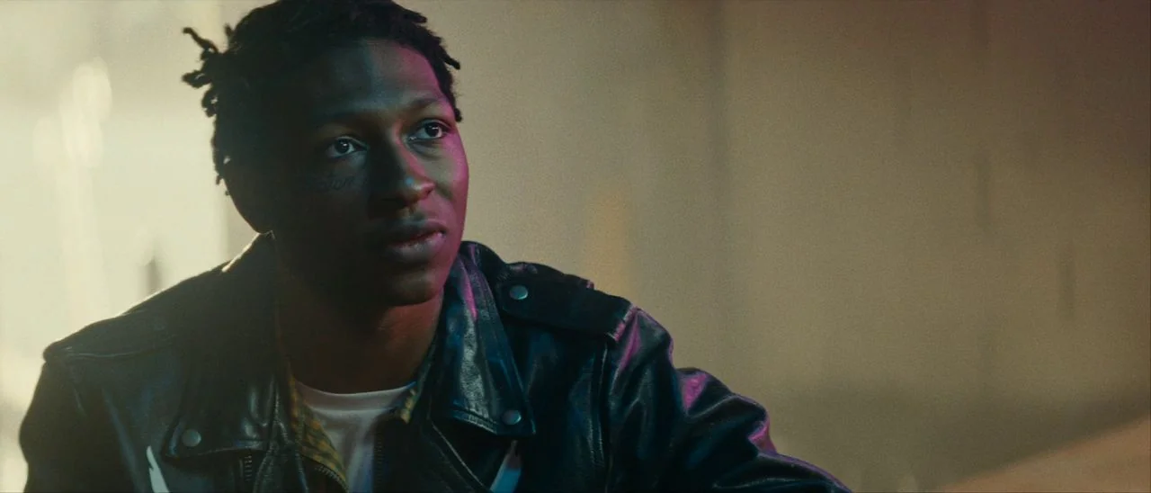 Archenemy (2020) movie still: medium — An interior space is cast in moody shadows with soft colored light. A young man with dreadlocks and a leather…