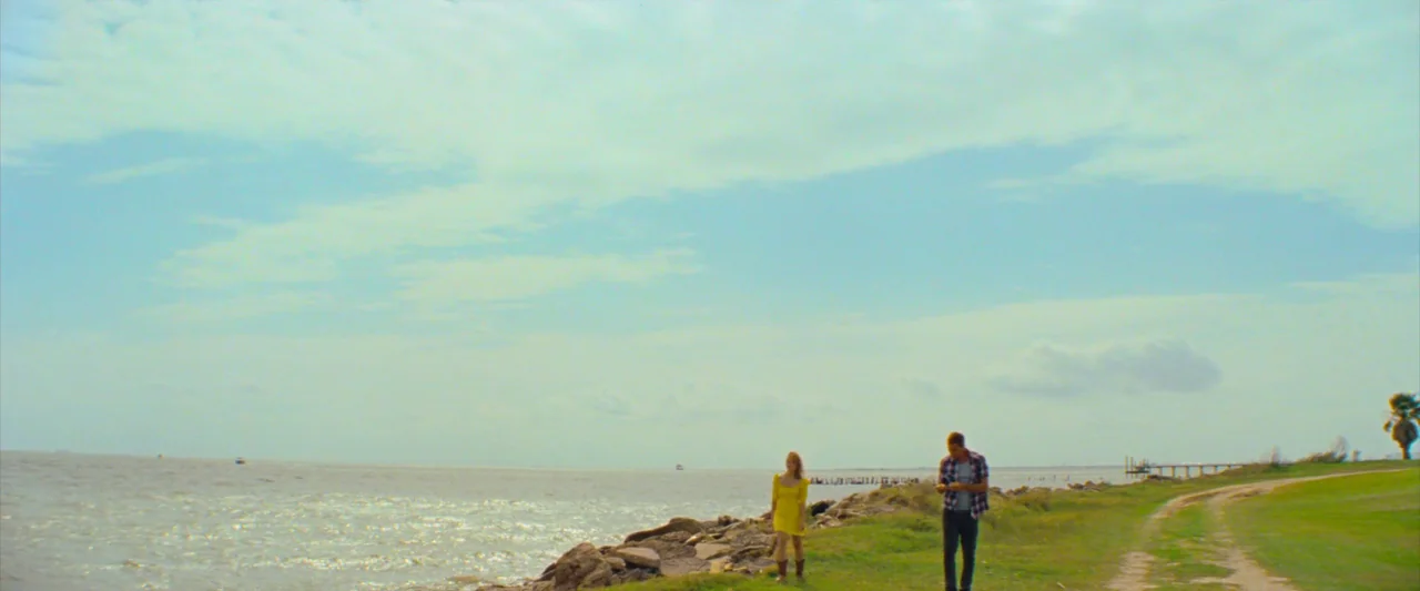 Red Rocket (2021) movie still: wide — A coastline with a rocky shore and a grassy path beside the water. A man and woman walk alongside each other…