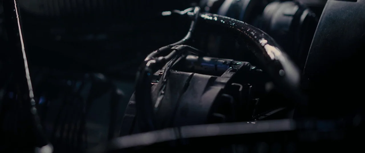 Titane (2021) movie still: closeup — A close-up view of a wet car engine interior with visible wiring and machinery. Metal components are coated in…