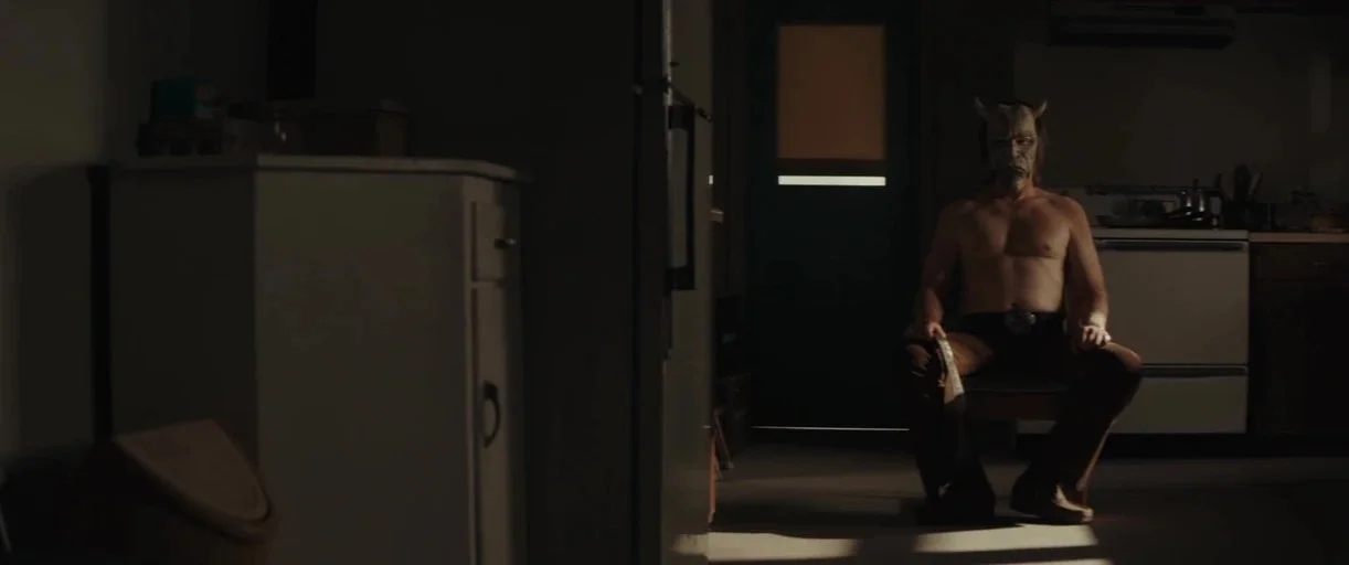 The Black Phone (2021) movie still: fullbody — A dimly lit room with a white stove and cabinets in the background. A man wearing a devil mask sits…