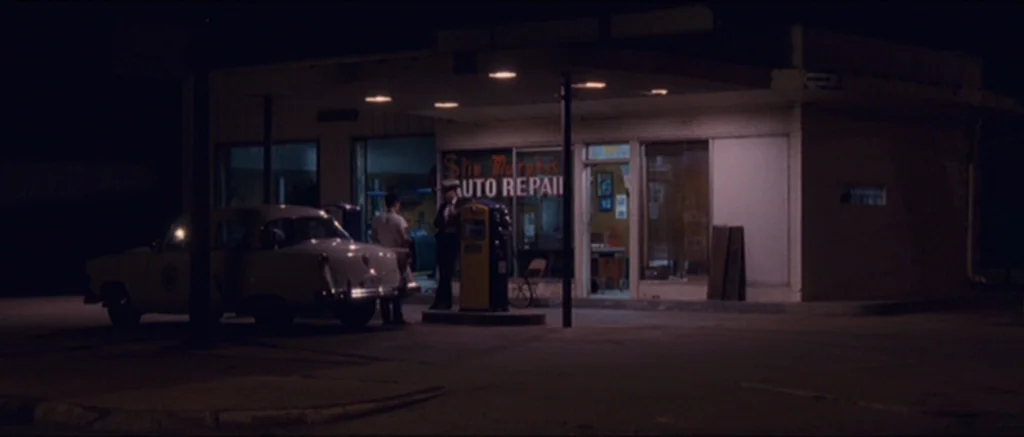 The Killer Inside Me (2010) movie still: wide — A small auto repair garage with a flat roof sits at night. Two people stand near a vintage yellow…