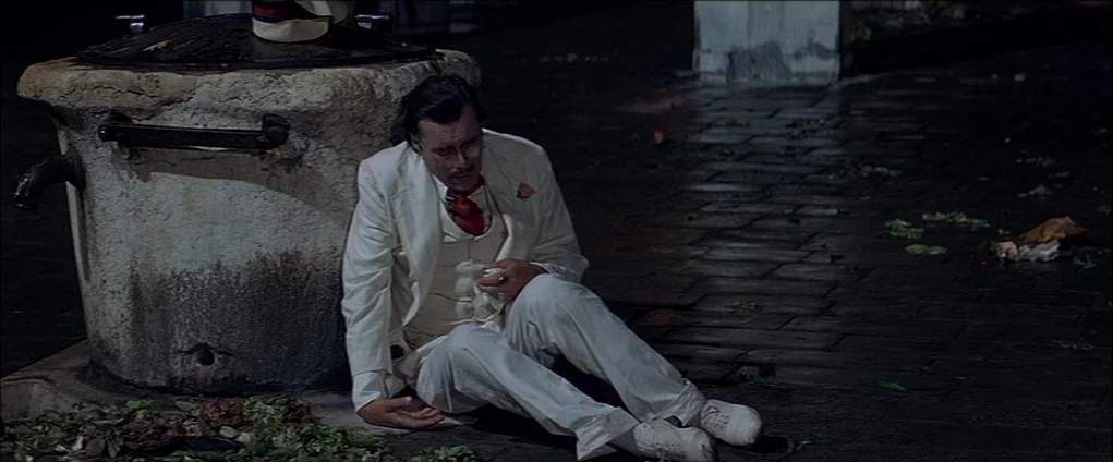 Death In Venice (1971) movie still: fullbody — A dark, paved square at night with a stone well feature. A man in a white suit sits slumped against…