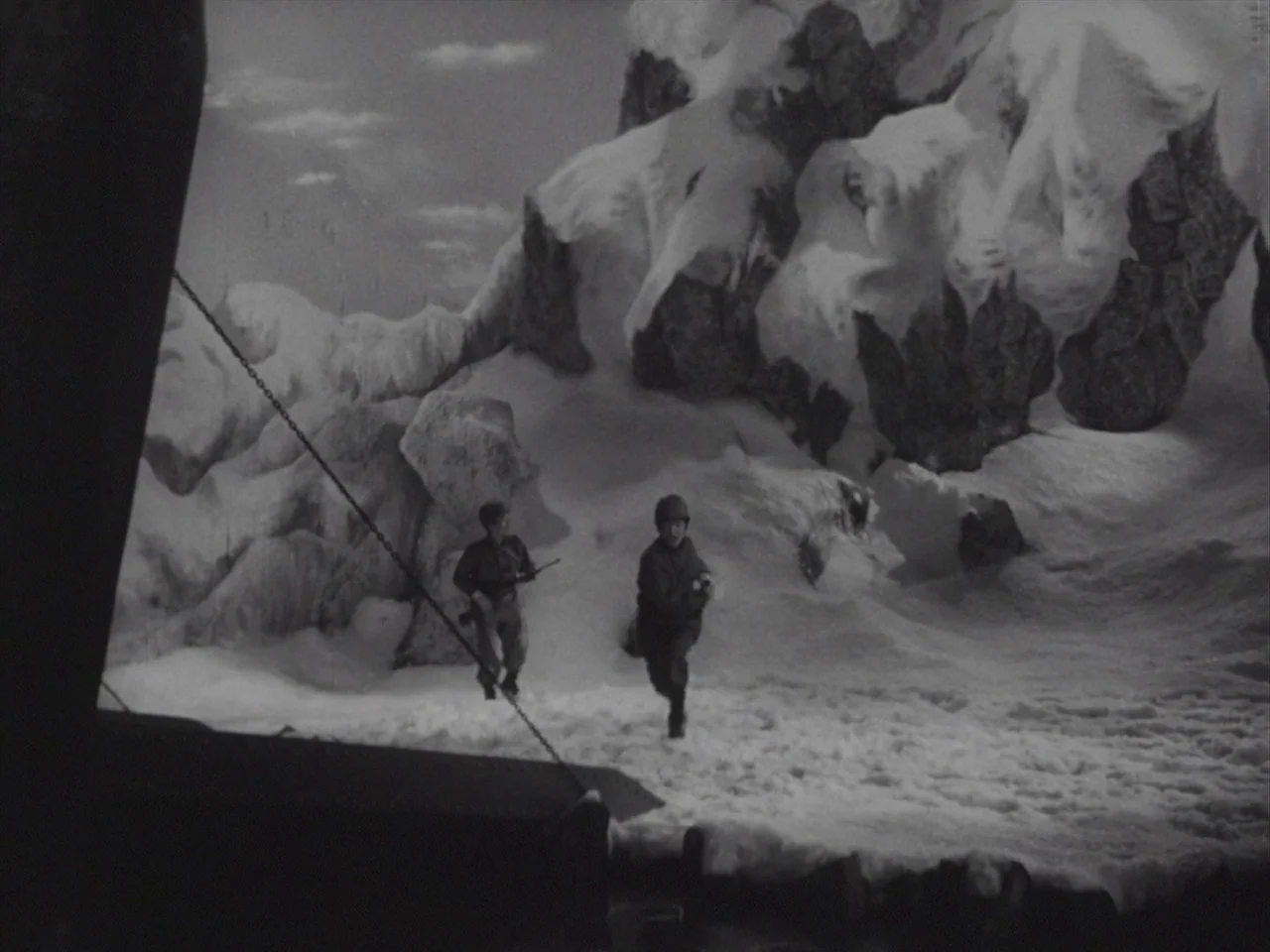 Godzilla Raids Again (1955) movie still: wide — Two soldiers walk across a vast, snow-covered mountain slope beneath a cloudy sky. The soldiers move…