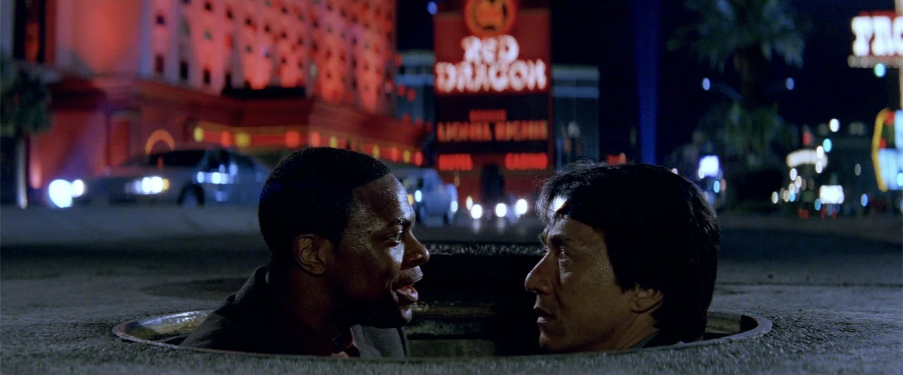 Rush Hour 2 (2001) movie still: medium — A manhole opening at night with an illuminated Las Vegas casino background. Two men emerge from the ground…