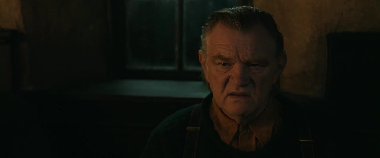 The Banshees of Inisherin (2022) movie still: closeup — A dark, rustic interior features a man positioned in front of a small window. The man looks…