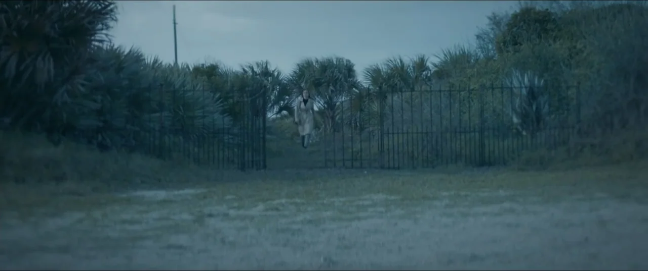 Offseason (2022) movie still: wide — A dark metal gate stands open in a grassy area surrounded by dense palm trees. A woman in a tan coat walks…