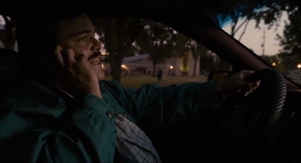 Bernie (2011) movie still: medium — A man drives a car at dusk with blurred suburban trees visible outside. He holds a cell phone to his ear while…