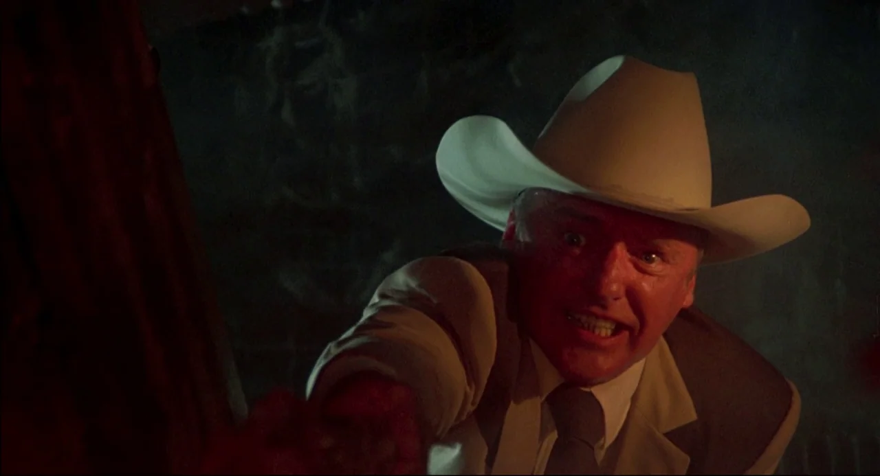 The Texas Chainsaw Massacre 2 (1986) movie still: closeup — A man in a tan suit and cowboy hat stands in a dark space. He reaches his hand toward…