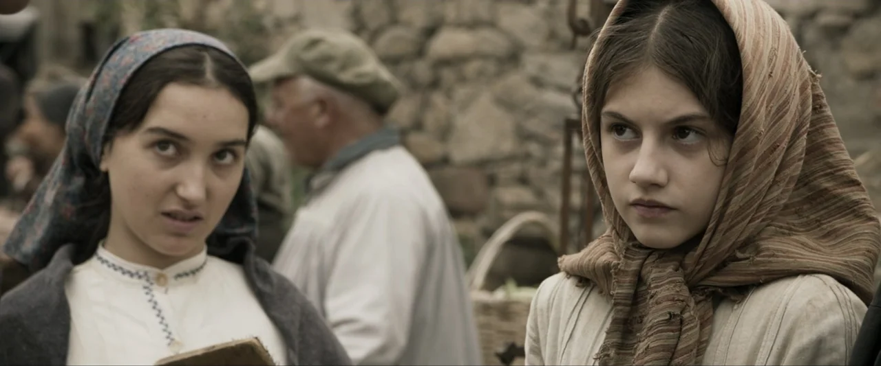 Fatima (2020) movie still: medium — A rustic stone village setting features people gathered in an outdoor marketplace. Two young women in…