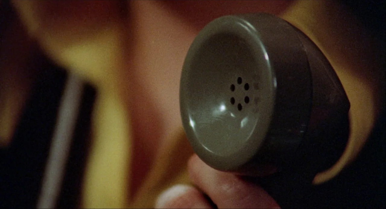 Black Christmas (1974) movie still: closeup — A close view shows a person holding a vintage plastic telephone receiver in their hand. The background…