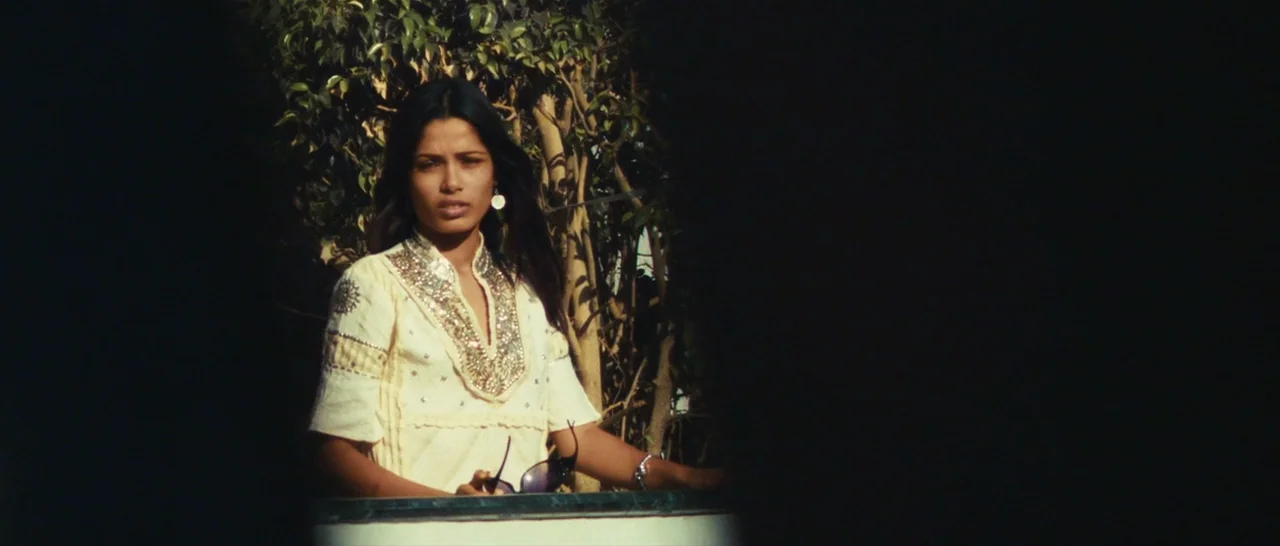 Slumdog Millionaire (2008) movie still: medium — A woman stands on an outdoor balcony framed by dense dark green foliage behind. She looks forward…