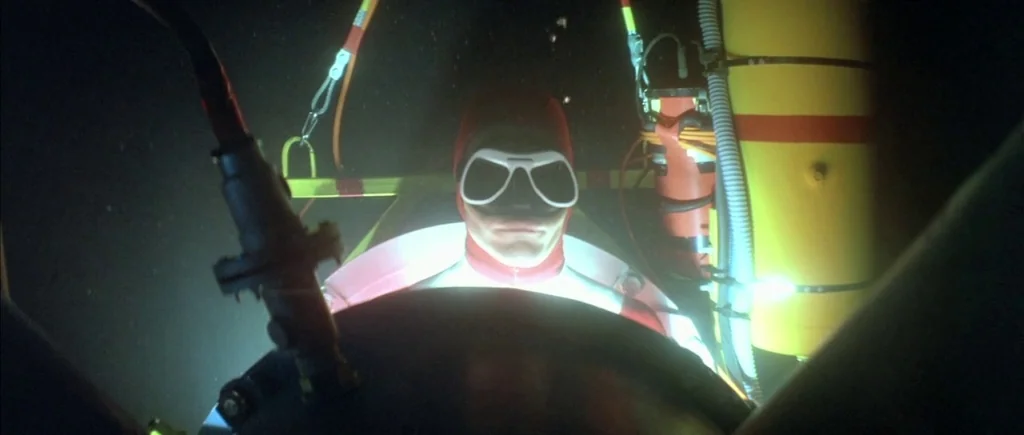 The Big Blue (1988) movie still: closeup — A dark, cramped interior of a small underwater vessel shows mechanical equipment and wires. A pilot in a…