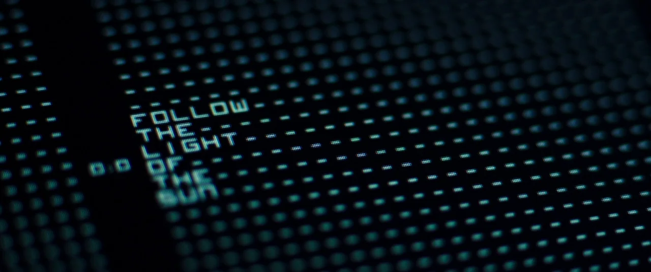 Assassin’s Creed (2016) movie still: closeup — A dark computer screen displays a glowing grid of blue pixels and text characters. The words follow…