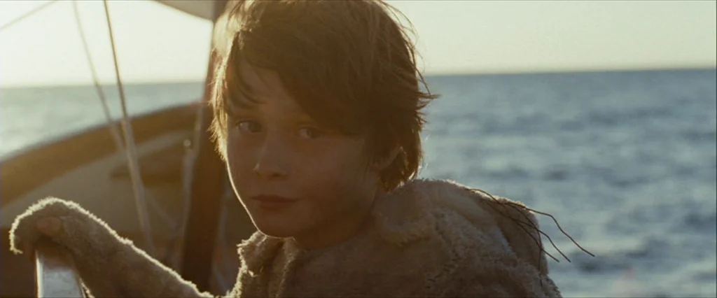 Where The Wild Things Are (2009) movie still: closeup — A boy stands on a boat deck against a backdrop of open ocean water. The late afternoon sun…
