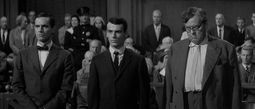 Compulsion (1959) movie still: medium — A formal interior space with dark wooden paneling and a crowd of seated observers. Three men stand in a line…