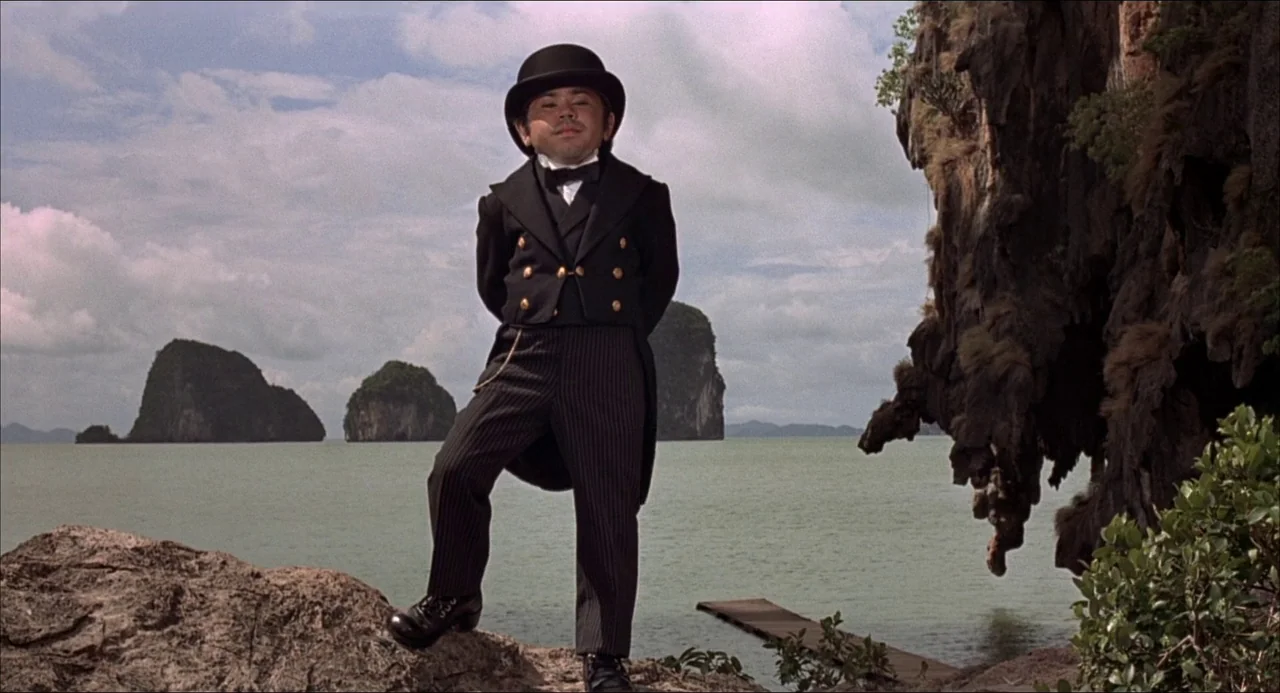 The Man With the Golden Gun (1974) movie still: fullbody — A man in a formal tuxedo stands on a rocky outcrop overlooking calm water. Distant…