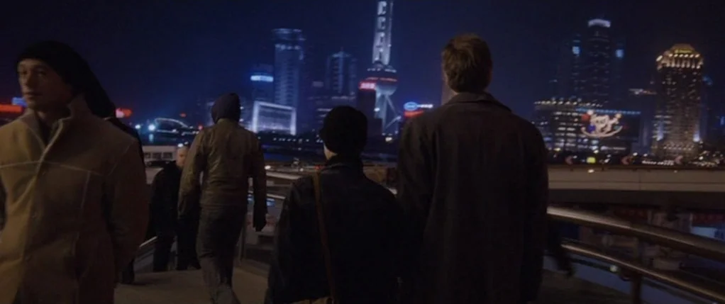 Code 46 movie still: medium — A night view of the Shanghai skyline across the water from an elevated walkway. Four figures walk away from the camera…
