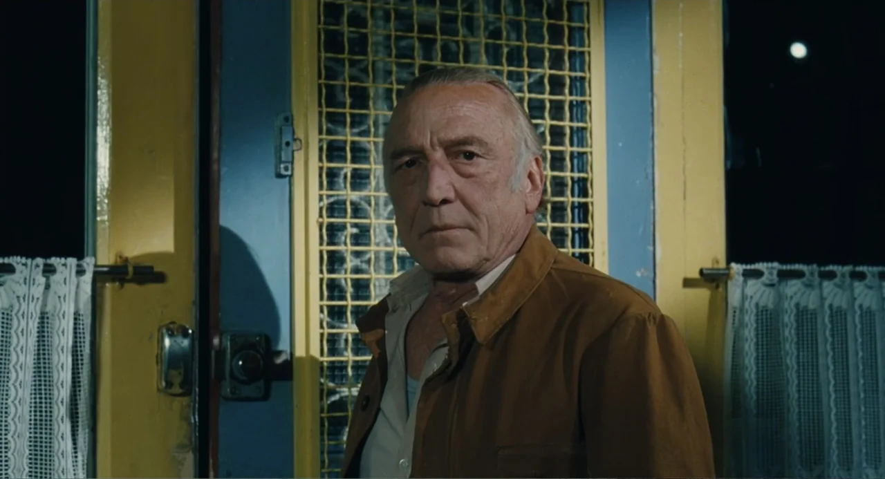 Le Havre (2011) movie still: medium — A man stands in front of a yellow door with a wire mesh screen. He remains stationary and faces the camera…