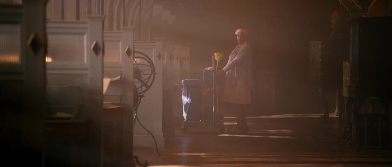Paddington (2014) movie still: wide — A quiet library interior features rows of wooden study carrels bathed in hazy light. A cleaning staff member…