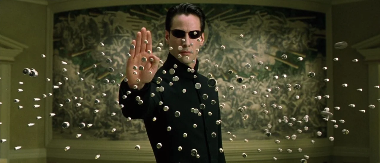 The Matrix Reloaded (2003) movie still: medium — A man stands in a hallway in front of an intricate wall tapestry. Dozens of metallic bullets hover…