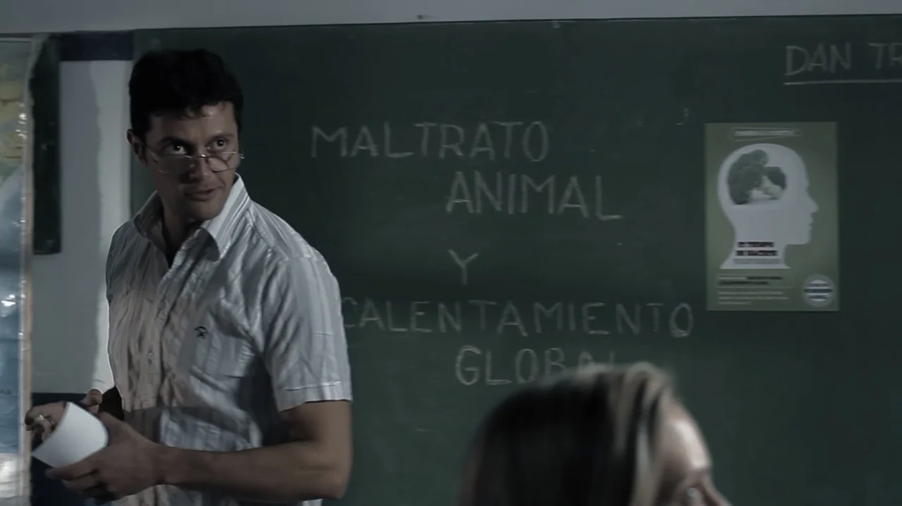 Still Life (2013) movie still: medium — A man stands before a dark green chalkboard with white hand-written text in Spanish. He looks toward the…