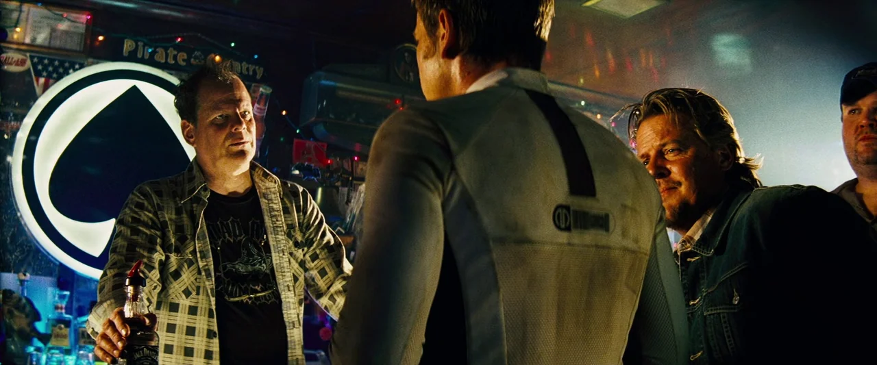 The Island (2005) movie still: medium — A dimly lit bar setting with a large spade-shaped neon sign mounted on the wall. Three men occupy the…