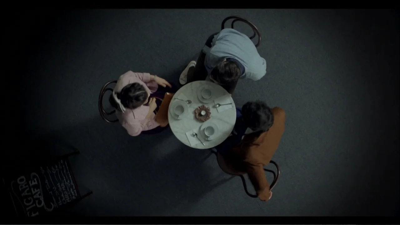 Six by Sondheim (2013) movie still: wide — A top-down view of three people seated at a small marble table indoors. They are gathered around empty…