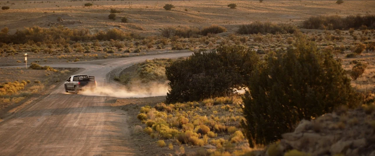 The Marksman (2021) movie still: wide — An arid desert landscape features a winding dirt road surrounded by low scrub brush. A dark pickup truck…