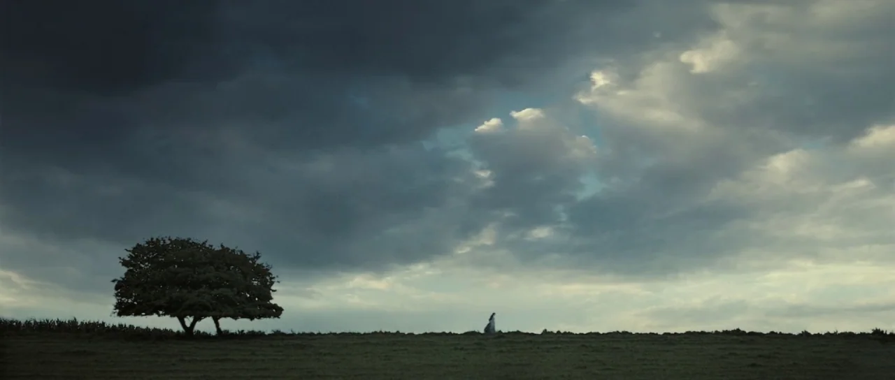 Pride & Prejudice (2005) movie still: wide — A vast, grassy field stretches under a heavy, overcast, dark gray storm sky. A lone person in a dress…