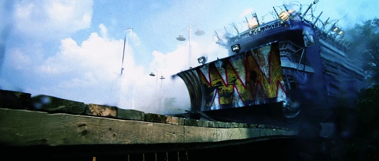 Dawn of the Dead (2004) movie still: wide — A heavy-duty armored vehicle with a jagged-toothed plow drives across a wooden bridge. Barbed wire wraps…