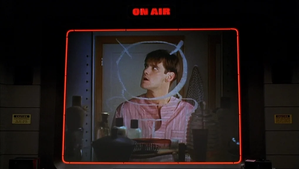 The Truman Show movie still: medium — A dark, industrial-style control room surrounds a large monitor showing a man inside a bathroom. The man…