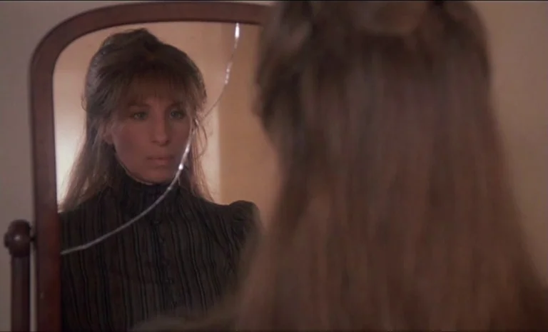 Yentl (1983) movie still: medium — A woman looks into a vintage oval mirror with a distinct crack in it. The frame shows her reflection from behind…