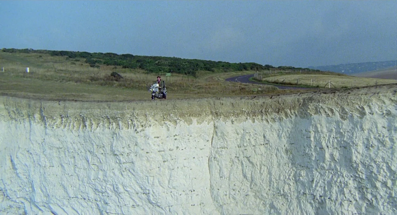 Quadrophenia (1979) movie still: wide — A man sits on a motorcycle at the edge of a tall white limestone cliff. Green hilly terrain and a distant…