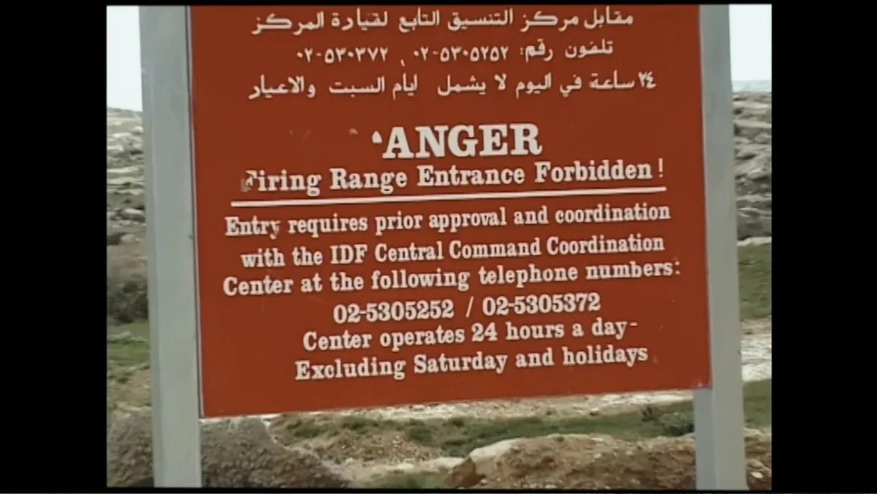 No Other Land (2024) movie still: closeup — A rectangular metal sign stands in a rocky, undeveloped outdoor field. The sign displays printed text in…