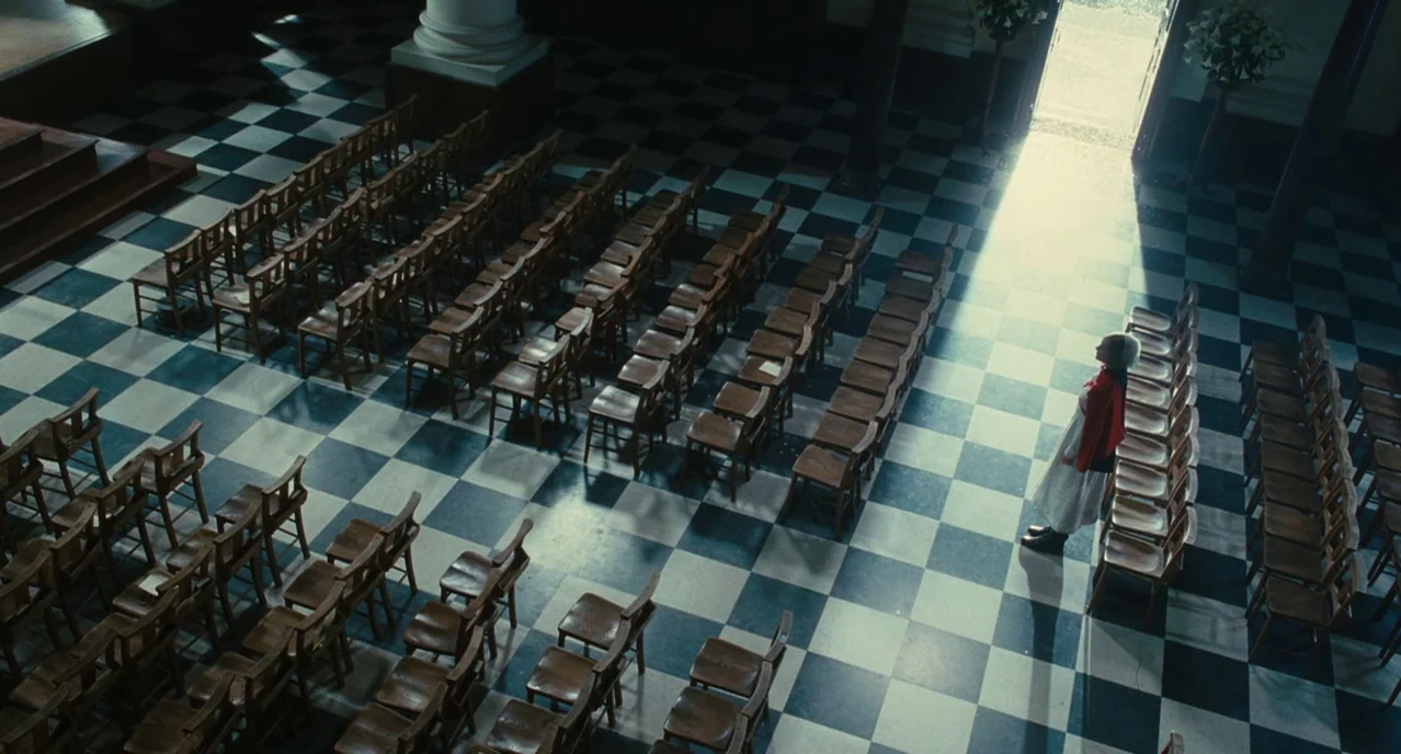Atonement (2007) movie still: wide — A spacious room features a distinct black and white checkered tile floor and many wooden chairs. A lone woman…