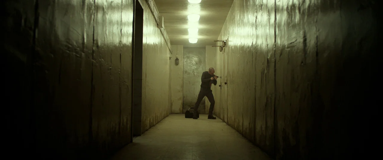 Split (2016) movie still: wide — A narrow, sterile hallway with textured, painted walls and a row of ceiling lights. A bald man stands alone, using…