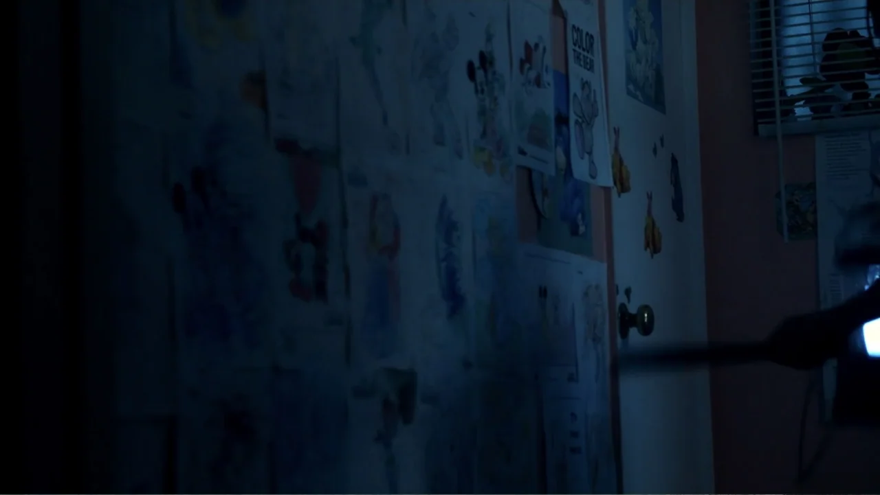 Abandoned Dead (2015) movie still: medium — A dark bedroom interior features a wall plastered with multiple children's drawings and posters. A…