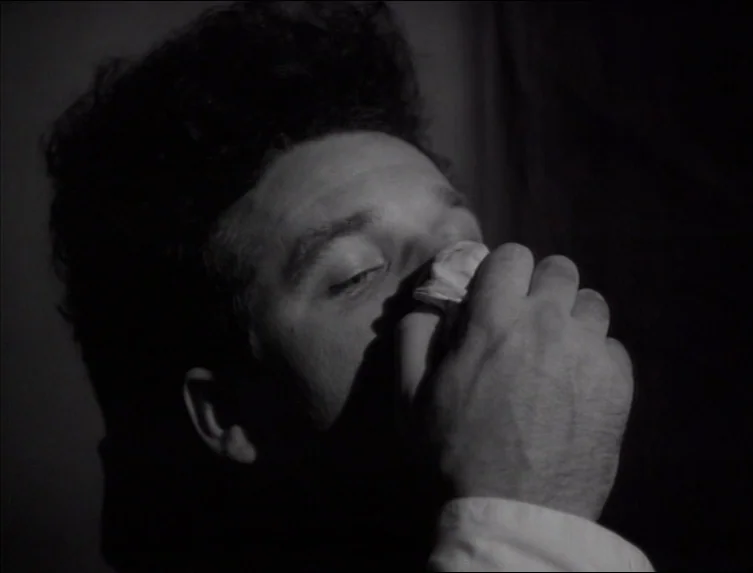 Eraserhead (1977) movie still: closeup — A dark indoor space with minimal background detail visible in the shadows. A man presses a white folded…