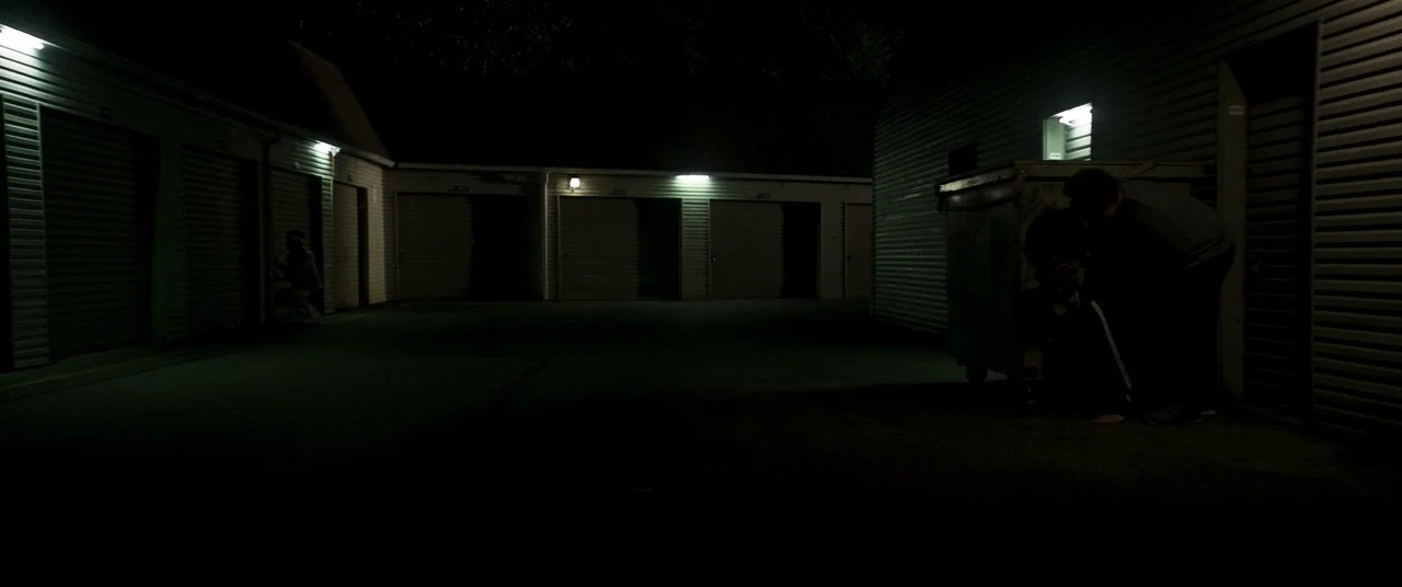 Summer of 84 (2018) movie still: wide — A dark, exterior storage unit facility with rows of metal garage doors at night. Two figures crouch low…