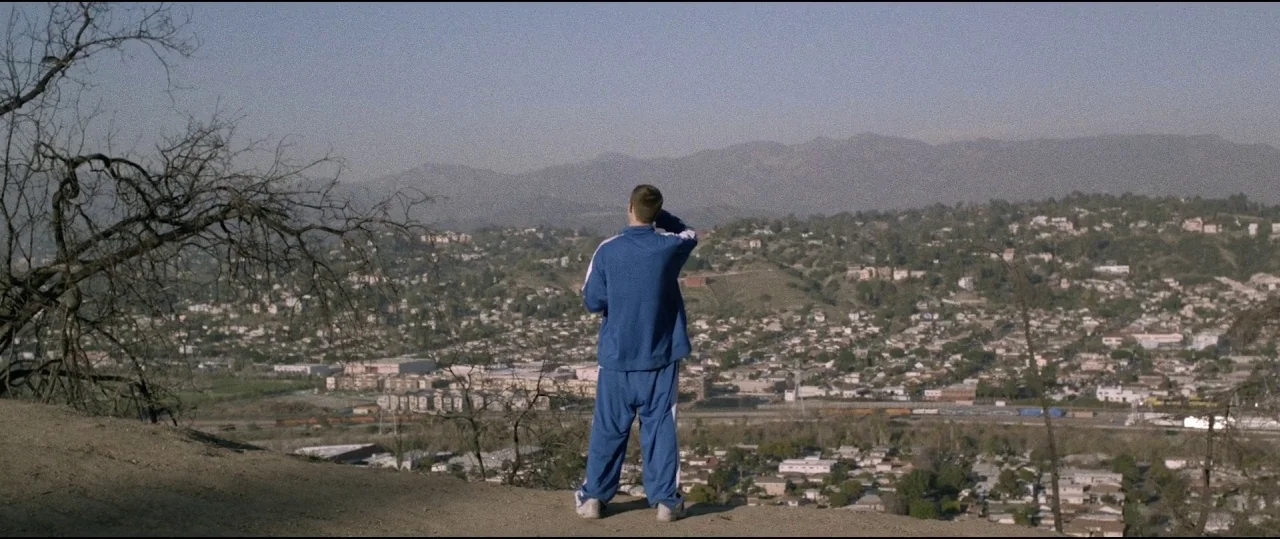 Lowlife (2017) movie still: wide — A dry, dusty hilltop offers a panoramic view over a densely packed residential valley. A young man wearing a blue…