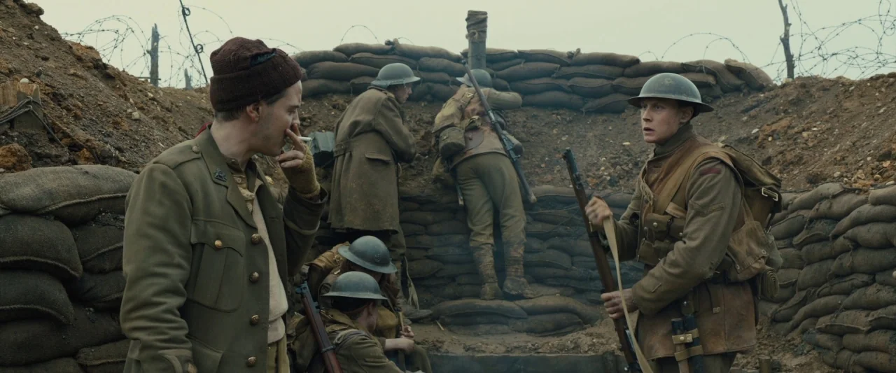 1917 movie still: medium — A muddy, narrow military fortification features stacked sandbags lining the earthen walls. Five soldiers in…