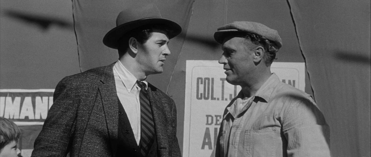 The Tarnished Angels (1957) movie still: medium — An outdoor political rally features posters with text displayed on a large curtain backdrop. Two…