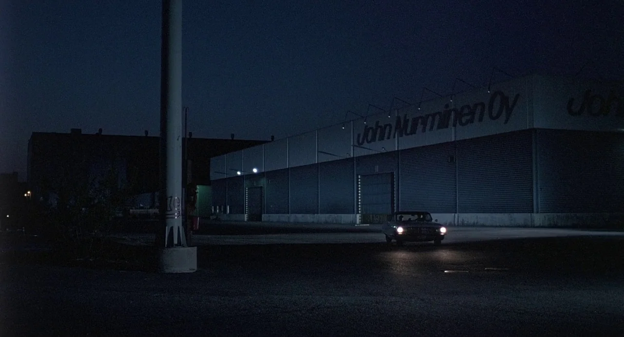 Ariel (1988) movie still: wide — A large warehouse exterior with John Nurminen Oy branding occupies the right side of frame. A car with illuminated…